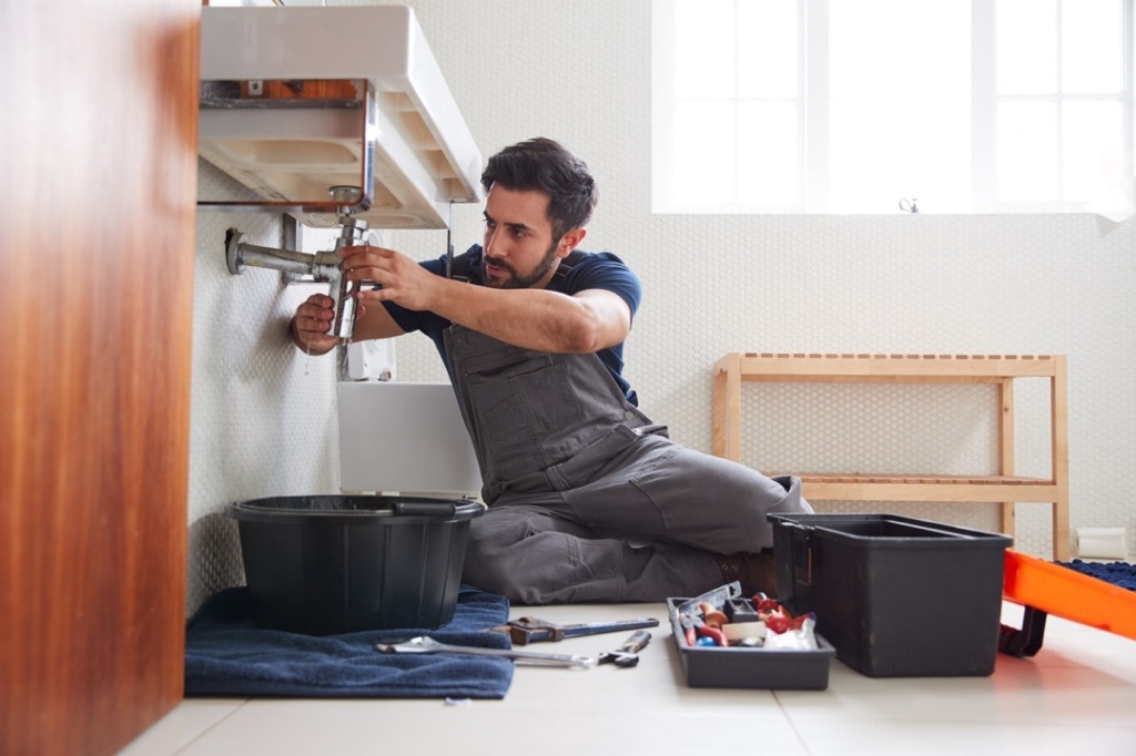 Person wearing coveralls sitting on a floor fixing a pipe under a sink with tools scattered around them